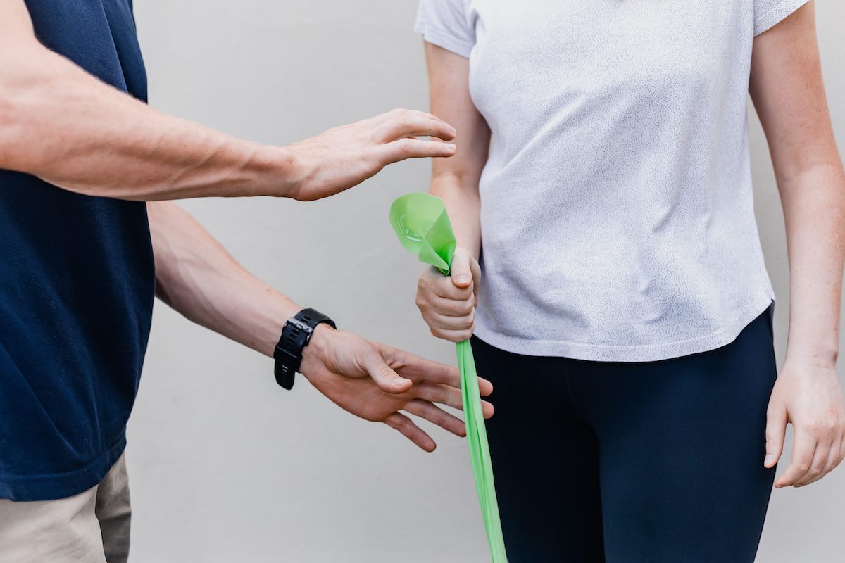 Restorative Physical Therapy. A physical therapist shows a patient an exercise where their arm is bent at the elbow and they are holding a stretchy band anchored on the floor.