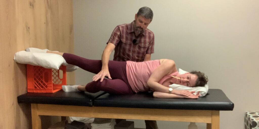 Postural Restoration PRI Retraining. A patient lies on their side with their bottom leg bent and top leg straight and a wooden splint between their upper and lower teeth. The physical therapist is holding the knee of the patient's bottom leg, their top leg is supported by a milk crate.
